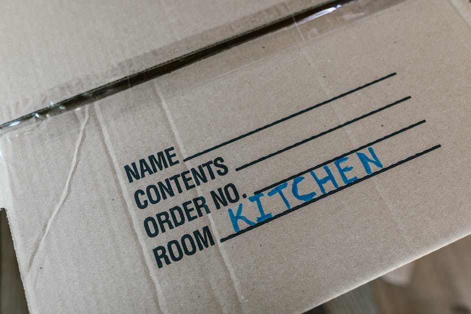 Close-up view of a cardboard moving box with a white and blue label indicating its contents in black text, including sections for name, contents, order number, and room. The room is marked as 'KITCHEN' in blue handwritten letters. The box is sealed with transparent packing tape and appears to be part of a home relocation or packing process. The background suggests an indoor environment, possibly a hallway or entryway, with additional boxes or packing materials partially visible. This image illustrates packing and moving procedures managed by Man and Van Kew as part of their removals service, capturing the organized aspect of furniture transport and home relocation logistics.