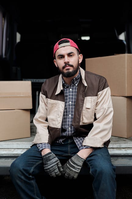 A man wearing a white T-shirt and jeans is lifting a cardboard box onto a stack of similar boxes near the open rear doors of a white moving van, which is parked on the street outside a residential pro