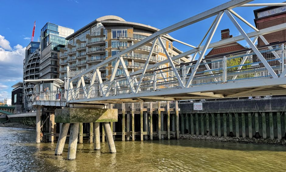 A pedestrian footbridge with white metal railings and support beams crosses over a river in an urban setting, with modern multi-storey residential and commercial buildings featuring large glass window