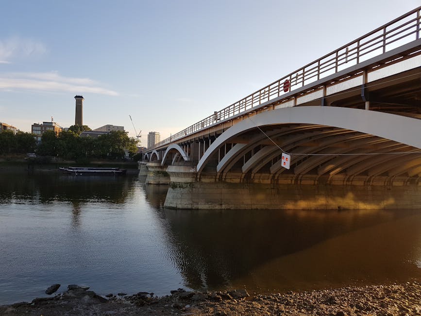 A pedestrian footbridge with white metal railings and support beams crosses over a river in an urban setting, with modern multi-storey residential and commercial buildings featuring large glass window
