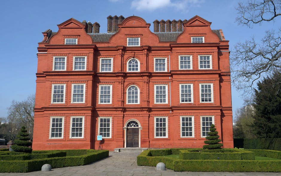 A large, historic red brick building with three stories, featuring an ornate, curved roofline and numerous symmetrically arranged white-framed sash windows. The entrance has a dark wooden door with a semi-circular window above it, accessed by a small set of stone steps. The building is set in a landscaped area with well-maintained green hedges, including low, rectangular hedges and two small, sculpted conical topiary trees flanking the pathway leading to the door. The paved pathway is lined with stone bollards on either side. The surrounding environment includes leafless trees and a clear blue sky, indicating a bright, sunny day. The scene represents a typical setting for house removals or home relocation services in an area like Kew, with the building visible from the front, and no moving activity or furniture present in this particular image. Man and Van Kew’s removals team may be involved in transporting household items, as suggested by the building’s prominent and inviting facade, suitable for a property undergoing relocation or packing during a move.