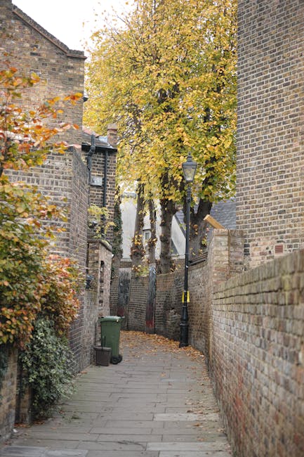 A narrow cobbled alleyway lined with brick buildings on both sides, with a green wheelie bin positioned on the left pavement and a black street lamp attached to a nearby brick wall. A large deciduous tree with yellow-orange leaves extends upward at the end of the alley, partially covering the view beyond. The scene appears to be part of a residential area, with a slight incline visible. The alley, which is part of a home relocation or furniture transport process, demonstrates confined access often encountered during house removals, with the texture of the bricks and pavement clearly visible, emphasizing the tight space for loading or unloading household items. The muted daylight and autumn foliage highlight the practical challenges addressed by professional removals services such as Man and Van Kew.