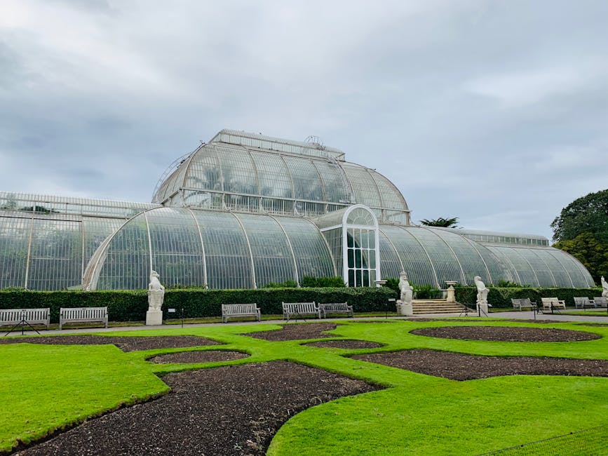 A large Victorian-style glass greenhouse with a domed roof and an arched central entrance, situated in the gardens of Kew Gardens. The greenhouse is constructed from metal and glass panels, with a pathway leading up to the entrance, flanked by decorative stone statues. In the foreground, well-maintained lawns with flower beds and winding gravel paths are visible, along with several wooden benches placed along the garden's edge. The scene is captured during daylight under a cloudy sky, with the lush greenery of the garden surrounding the greenhouse. The image reflects a peaceful outdoor environment characteristic of a historic botanical estate, and is relevant to house removals and moving services related to property upgrades or relocations near iconic landmarks such as Kew Gardens, as provided by Man and Van Kew.