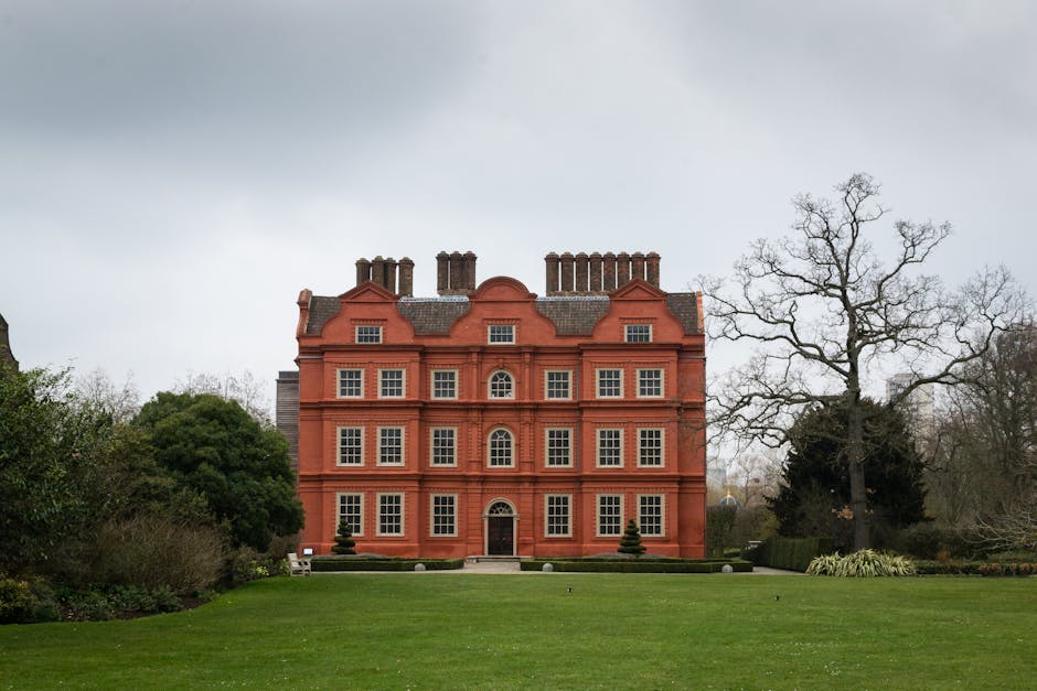 A large, multi-storey building constructed of red brick, with a symmetrical facade featuring numerous white-framed sash windows arranged in a grid pattern. The building has a central arched doorway and decorative brickwork accents. In front of the building, a well-maintained lawn with short grass extends to the edge of the image, with a few small, neatly trimmed conifer bushes near the entrance. To the right of the building, a tall, leafless tree stands alongside other trees and shrubs, indicating a winter or early spring season. The sky above is overcast with grey clouds, creating diffuse lighting across the scene. This image is part of a house removal or relocation context, possibly captured during a packing or loading process involving Man and Van Kew, highlighting the transportation of domestic furniture or large items in preparation for a home move near the Kew Gardens area.