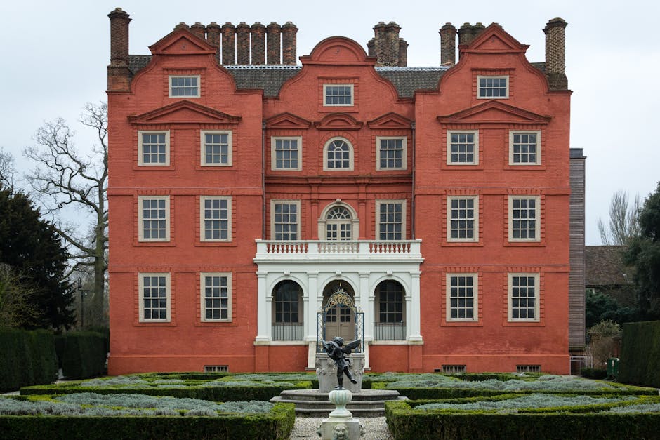 A large Victorian-style glass greenhouse with a domed roof and an arched central entrance, situated in the gardens of Kew Gardens. The greenhouse is constructed from metal and glass panels, with a pathway leading up to the entrance, flanked by decorative stone statues. In the foreground, well-maintained lawns with flower beds and winding gravel paths are visible, along with several wooden benches placed along the garden's edge. The scene is captured during daylight under a cloudy sky, with the lush greenery of the garden surrounding the greenhouse. The image reflects a peaceful outdoor environment characteristic of a historic botanical estate, and is relevant to house removals and moving services related to property upgrades or relocations near iconic landmarks such as Kew Gardens, as provided by Man and Van Kew.