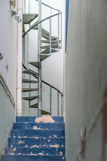 A narrow, indoor staircase with a low ceiling, featuring a blue, worn and chipped wooden step surface, leading upward to a small landing. To the left side, there are white plumbing pipes running beside the wall, with a small, mounted white spotlight visible near the top of the stairs. A metal spiral staircase with open risers and a black handrail is positioned in the corner, connecting to the upper floor. The surrounding walls are plain and painted in neutral colours, with some visible signs of wear and minor damage. The setting appears to be inside a residential property, where furniture or belongings may need to be carefully maneuvered to accommodate the low ceiling height, as part of a house relocation process. This environment aligns with challenges faced during furniture transport and packing in tight internal spaces. Man and Van Kew, a professional removal service, often handles such detailed home moving logistics.