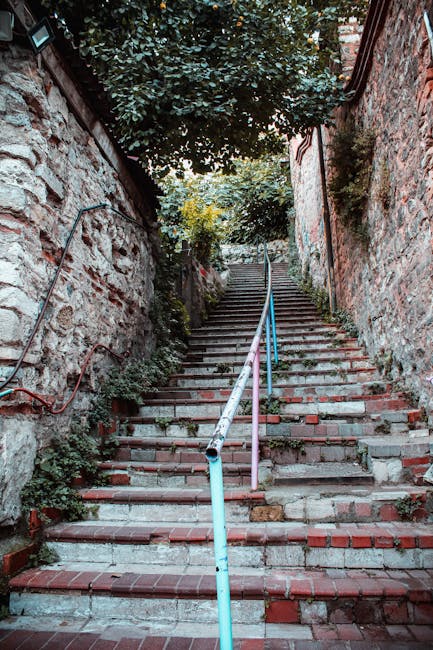 A long flight of narrow, uneven outdoor stairs made of concrete and brick leading upward between two brick and stone walls. The stairs are bordered by metal handrails painted light blue, with some sections showing signs of rust and wear. At the top, there is a lush green tree and sunlight filtering through the leaves, suggesting a residential area with limited ceiling height. The steps are cluttered with a few small cardboard boxes, some wrapped in plastic, and a lightweight fabric moving blanket, indicating a home relocation process. The surrounding environment appears aged, with patches of moss and weeds growing between the cracks, and electrical wiring visible along one of the walls. This scene depicts the challenging access often encountered in house removals when delivering or removing furniture and boxes from properties with low ceiling restrictions, making careful loading and maneuvering necessary, as managed by companies like Man and Van Kew.