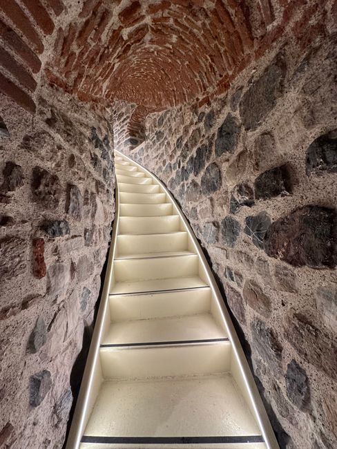 A narrow, indoor staircase with a low ceiling, featuring a blue, worn and chipped wooden step surface, leading upward to a small landing. To the left side, there are white plumbing pipes running beside the wall, with a small, mounted white spotlight visible near the top of the stairs. A metal spiral staircase with open risers and a black handrail is positioned in the corner, connecting to the upper floor. The surrounding walls are plain and painted in neutral colours, with some visible signs of wear and minor damage. The setting appears to be inside a residential property, where furniture or belongings may need to be carefully maneuvered to accommodate the low ceiling height, as part of a house relocation process. This environment aligns with challenges faced during furniture transport and packing in tight internal spaces. Man and Van Kew, a professional removal service, often handles such detailed home moving logistics.