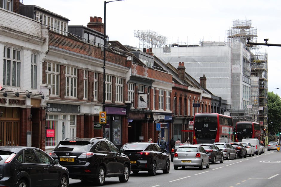 A street scene in an urban residential area during daytime, featuring a row of brick terraced houses with large front windows and traditional architectural details. In the foreground, several parked cars, including sedans and an SUV, line the curb alongside a busy pavement. Two red double-decker buses are in motion, one displaying route number 343. Some pedestrians are visible walking along the sidewalk near storefronts, and a shop with a sign that reads 'Candy' can be seen. To the right, a building under extensive scaffolding and wrapped in protective white sheeting suggests ongoing construction or renovation work. The street is well-lit with natural daylight, and the scene captures the typical environment for house removals and moving logistics in a city setting, emphasizing the urban infrastructure and parking considerations relevant to relocating furniture and household items. Man and Van Kew occasionally appears in context as a service provider operating within this environment.