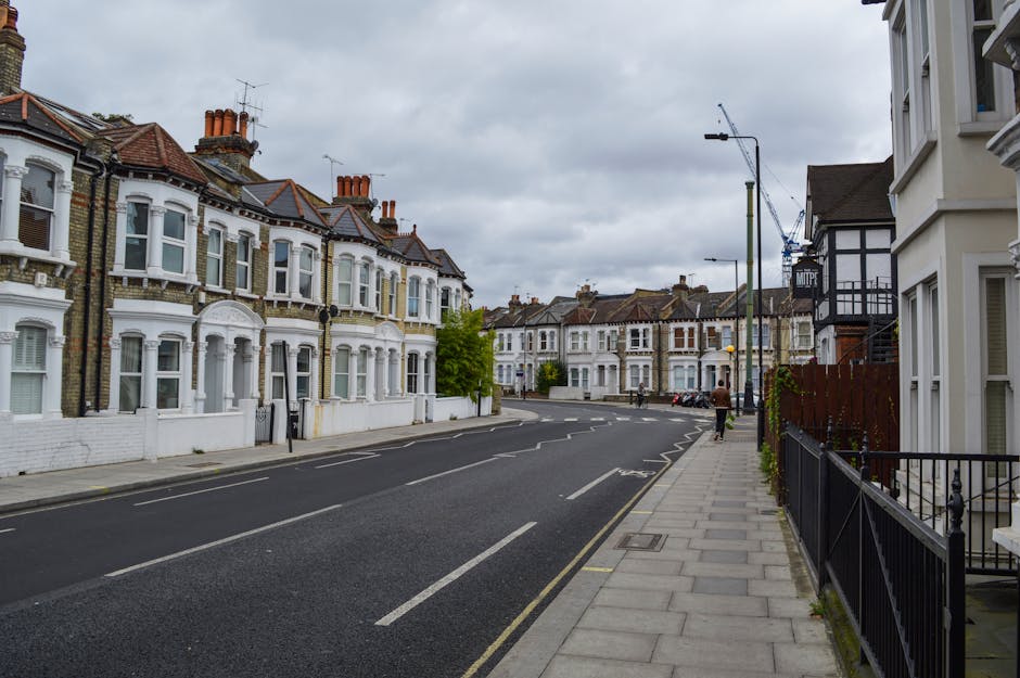 A wide view of a residential street showing the exterior of row houses with ornate bay windows, decorative brickwork, and tiled roofs under an overcast sky. The pavement runs alongside the houses, with a few pedestrians visible in the distance. On the right, a white building with black timber framing extends toward the sidewalk, partially obscured by a black metal fence. The street appears calm, with parked cars along the curb and no active loading activities currently happening. The scene reflects a typical UK street environment suitable for home relocation and furniture transport planning, such as those provided by Man and Van Kew, with the focus on architecture, street layout, and urban environment.