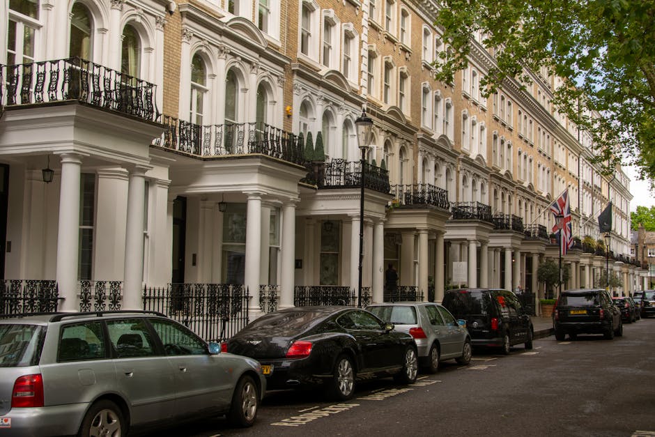 A street scene in an urban residential area during daytime, featuring a row of brick terraced houses with large front windows and traditional architectural details. In the foreground, several parked cars, including sedans and an SUV, line the curb alongside a busy pavement. Two red double-decker buses are in motion, one displaying route number 343. Some pedestrians are visible walking along the sidewalk near storefronts, and a shop with a sign that reads 'Candy' can be seen. To the right, a building under extensive scaffolding and wrapped in protective white sheeting suggests ongoing construction or renovation work. The street is well-lit with natural daylight, and the scene captures the typical environment for house removals and moving logistics in a city setting, emphasizing the urban infrastructure and parking considerations relevant to relocating furniture and household items. Man and Van Kew occasionally appears in context as a service provider operating within this environment.