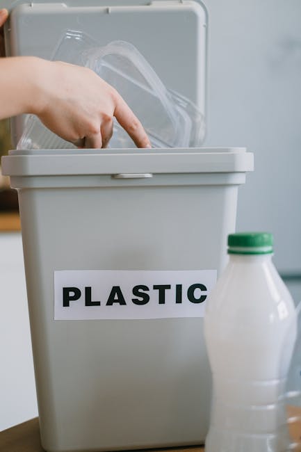 A close-up image showing a white plastic recycling bin with a black and white label reading 'PLASTIC' on the front. The lid of the bin is open, revealing transparent plastic packaging inside. In the foreground, a partially visible white plastic bottle with a green cap is placed on a surface. The background appears to be an indoor setting with a plain wall, typical of a home or property during a house removal or home relocation. This scene illustrates waste separation and disposal of plastic materials following a move, supported by Man and Van Kew’s removal services, emphasizing proper packing and disposal practices after a residential move.