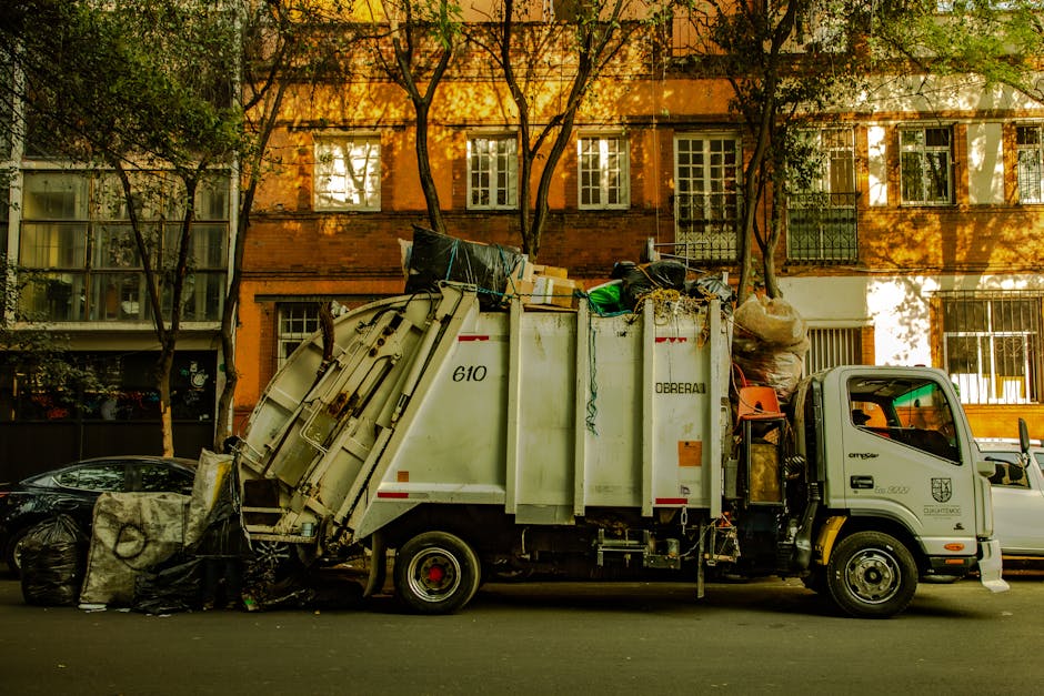 A white waste collection truck parked on a street in front of a multi-storey residential building with orange brick facade and white-framed windows. The truck, operated by Man and Van Kew, is loaded with various household items and waste materials including cardboard boxes, large bags of rubbish, plastic containers, and packing materials. The back of the truck is open, revealing a pile of items awaiting disposal, with some items extending onto the pavement. Nearby, a black car is partially visible, and the truck is positioned close to the curb, ready for collection as part of the home relocation or moving process. The scene is illuminated by natural daylight, with trees lining the street, casting shadows on the building and the vehicle, illustrating the logistics involved in residential waste disposal following a house move, as guided by local regulations in Kew.