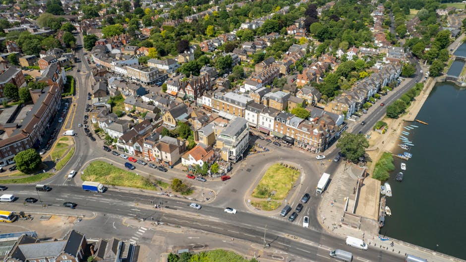An aerial view of a townscape featuring a mixture of terraced houses, shops, and commercial buildings along a main road that curves around a small green square. To the right, a river with a promenade and moored boats is visible, with trees lining the waterfront. The scene includes a busy road with cars, trucks, and buses moving through the area, and a parking area adjacent to the commercial buildings. Inside this urban environment, there are visible street furniture such as benches and lampposts, along with some parked vehicles near the shops. The overall setting showcases a typical town centre, suitable for home relocation or furniture transport services, with Man and Van Kew occasionally involved in the logistics of house removals and packing materials seen on-site. Bright afternoon lighting illuminates the scene, highlighting the detailed layout of streets, buildings, and the nearby waterway, emphasizing the logistical considerations for residential and commercial moving operations in accordance with Richmond upon Thames council removals permit rules for Kew.