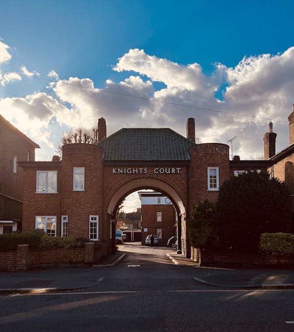 A brick residential building with a large arched entrance labeled 'KNIGHTS COURT' situated within a suburban street under a partly cloudy sky. The entrance leads to a parking area visible through the arch, where several cars are parked. On either side of the arch, there are small shrubbery and low brick walls bordering the pavement. The building features a tiled roof, multiple windows with white frames, and chimneys. The scene appears to be part of a residential complex in Kew, with the architectural style and surrounding environment characteristic of a suburban area in Richmond upon Thames. This location may be involved in house removals or residential logistics services, such as those offered by Man and Van Kew, particularly in relation to complying with local removals permit rules.