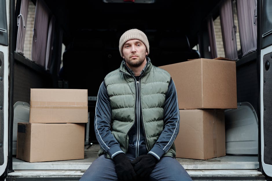 A man wearing a beige knit beanie, a green puffer vest over a navy blue jacket with white stripes on the sleeves, black gloves, and dark trousers, sits inside the open rear of a moving van. The van’s interior shows several cardboard boxes of various sizes, some sealed with packing tape, and some stacked on top of each other. The boxes are made of brown corrugated cardboard, and some are wrapped in clear plastic or fabric padding for protection during home relocation. The van is parked on a paved area, potentially outside a residential property, with the vehicle’s rear doors open to allow access to the packing and loading process. The scene depicts the process of furniture transport and packing during a house removal, with the van prepared for transportation of household items, aligning with professional moving services such as those offered by Man and Van Kew, specializing in removals and packing logistics.