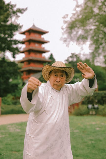 The image shows an elderly woman dressed in traditional white Asian clothing, standing outdoors in front of a multi-tiered pagoda with red and brown accents, surrounded by lush green trees and grass. She is wearing a wide-brimmed straw hat and has a focused expression on her face, with one hand raised in a greeting or gesture. In the background, there is a paved pathway and the atmosphere suggests a peaceful park or garden setting, possibly within a cultural or historical site. This scene may be associated with cultural themes such as traditional practices or heritage sites, related to house removals or relocation services that involve transport of belongings through scenic or heritage locations, as referenced on manandvankew.org.uk.