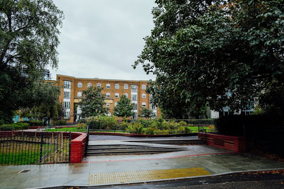 The image shows a residential exterior scene during daytime with a paved path leading to a staircase, which is part of a house removal and furniture transport plan for Kew Green terraces, KEW. The pathway is bordered by a black metal fence with red brick pillars on each side and is covered with a weather-resistant material. There are metal handrails on either side of the stairs to assist with the loading process. Behind the steps, a well-maintained garden area contains various shrubs and small flowering plants, suggesting it is part of a property being prepared for a house move. In the background, a mid-rise brick apartment building with multiple white-framed windows and balconies is visible, indicating urban living surroundings. Large leafy trees on both sides of the scene provide shade and greenery. The lighting is natural and overcast, typical of a cloudy day, making this setting suitable for a professional removals service such as Man and Van Kew, specializing in house removals and packing and moving logistics.