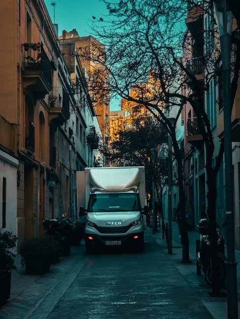 A white Iveco moving van parked on a narrow urban street, surrounded by residential buildings with balconies and large windows, some with potted plants. The van is positioned facing towards the viewer